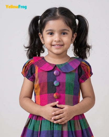 A young girl with dark hair in pigtails smiling, wearing a colorful plaid dress with pink, purple, green, and orange patterns. The YellowFrog brand logo is visible in the top left corner against a light gray background.