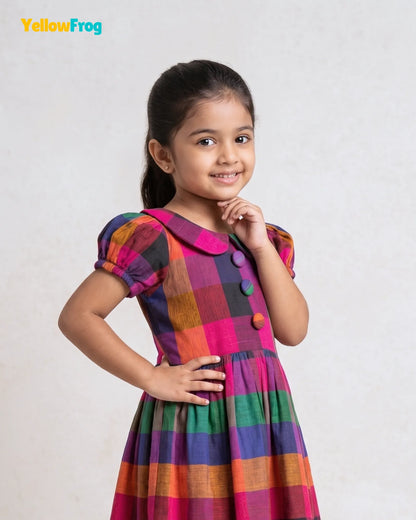 A young girl with dark hair tied back is wearing a colorful plaid dress with short puff sleeves and decorative buttons. She is posing against a light gray textured background, with the YellowFrog brand logo in the top left corner.