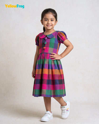 A young girl stands in a studio wearing a colorful plaid dress with puff sleeves, a round collar, and colorful buttons, paired with white sneakers. The YellowFrog logo is visible in the top left corner.