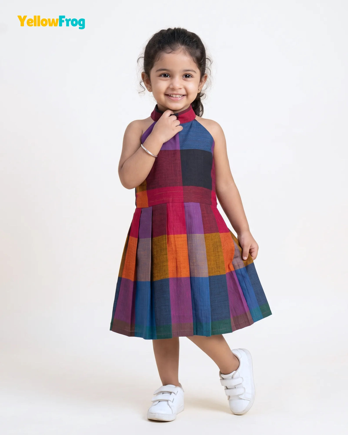 A young girl modeling a sleeveless halter-neck dress with a colorful patchwork plaid pattern, standing against a plain white studio background. The dress features blocks of red, purple, orange, blue, and green.