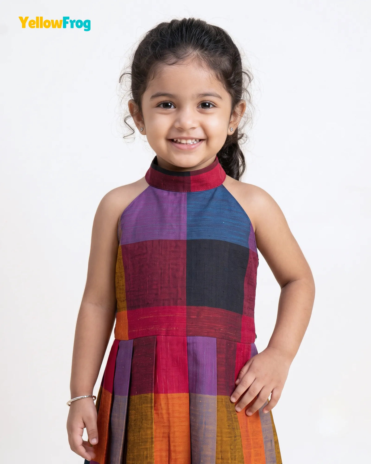 A young girl smiling and wearing a sleeveless halter-neck dress with a colorful patchwork pattern of purple, blue, red, black, orange, and yellow blocks, set against a plain white background with a YellowFrog logo in the top left corner.