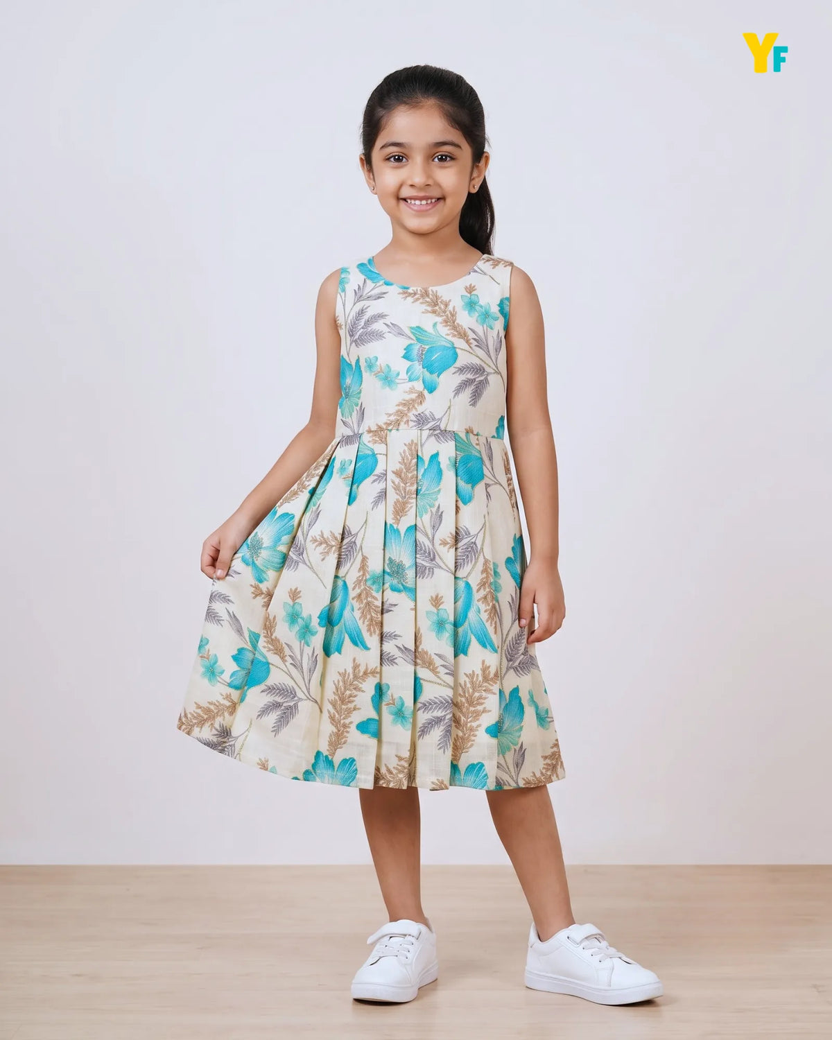 A young girl stands in a studio wearing a sleeveless pleated dress with a blue and beige floral print, paired with white sneakers. A small yellow crown emoji is visible in the top right corner.