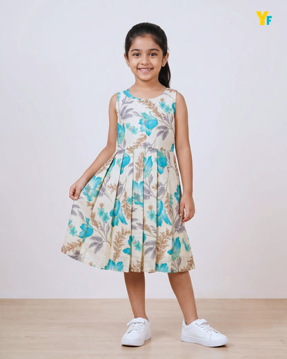 A young girl stands in a studio wearing a sleeveless pleated dress with a blue and beige floral print, paired with white sneakers. A small yellow crown emoji is visible in the top right corner.