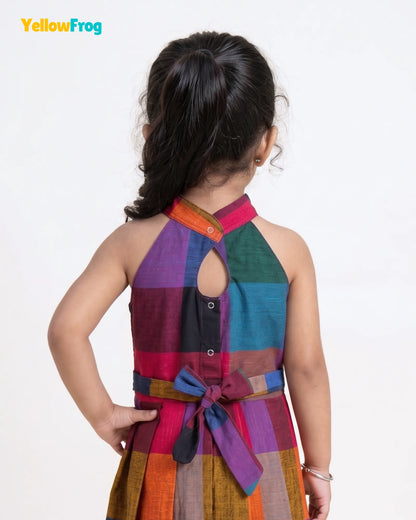 A young girl is shown from behind wearing a colorful patchwork halter dress with a tie belt at the waist. The dress features a mix of purple, green, blue, orange, and brown blocks.