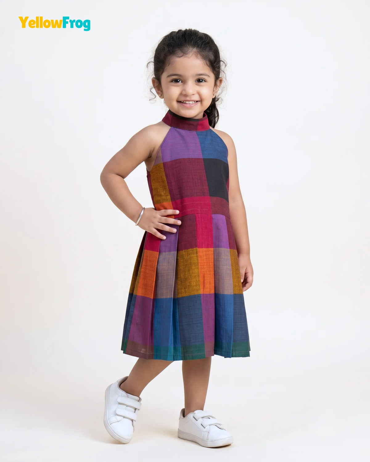 A young girl modeling a sleeveless halter-neck dress with a colorful patchwork plaid pattern, standing against a plain white studio background. The YellowFrog brand logo is visible in the top left corner.