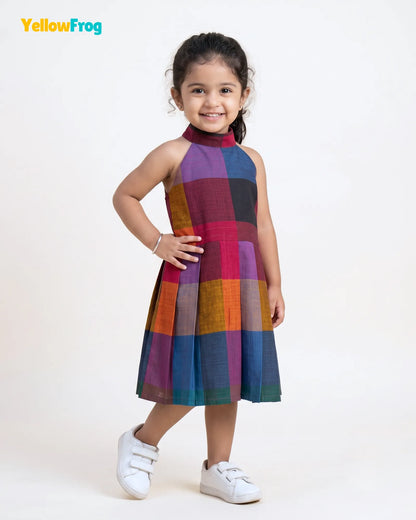 A young girl modeling a sleeveless halter-neck dress with a colorful patchwork plaid pattern, standing against a plain white studio background. The YellowFrog brand logo is visible in the top left corner.