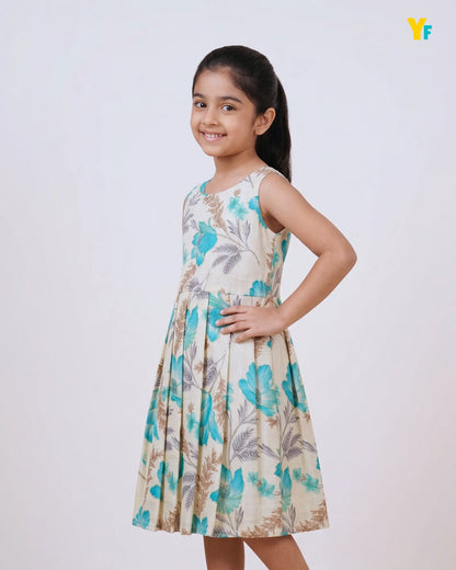 A young girl with her hair in a ponytail is wearing a sleeveless, pleated A-line dress with a blue and beige floral pattern, posing against a light gray studio background.