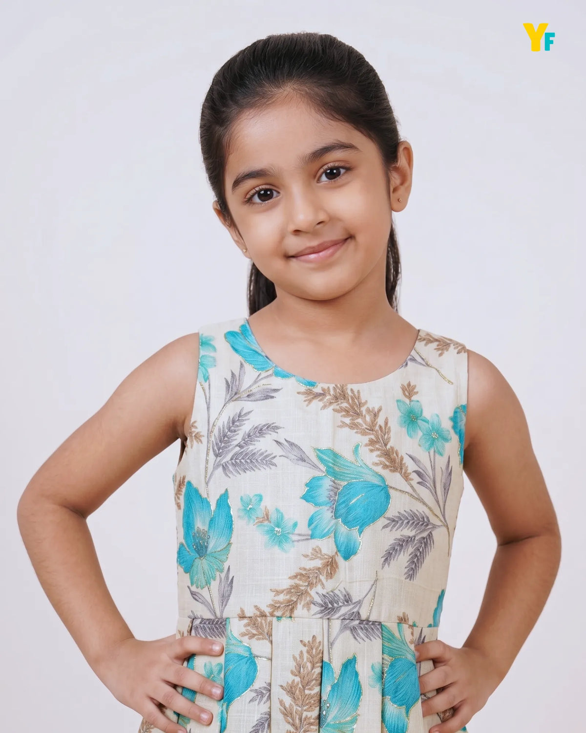 A young girl with dark hair tied back, wearing a sleeveless cream-colored dress with a blue and brown botanical pattern, posing with her hands on her hips against a light gray background.