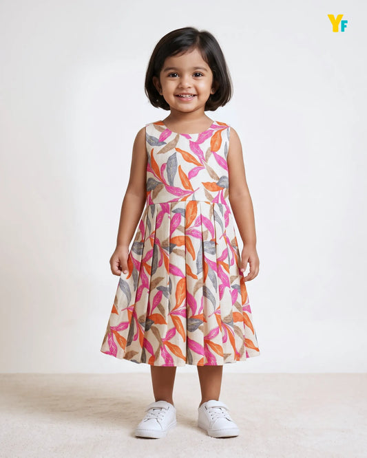 A young girl with short dark hair stands smiling in a sleeveless A-line dress with a colorful abstract leaf pattern on a cream background. She is wearing white sneakers against a clean, light off-white studio background.