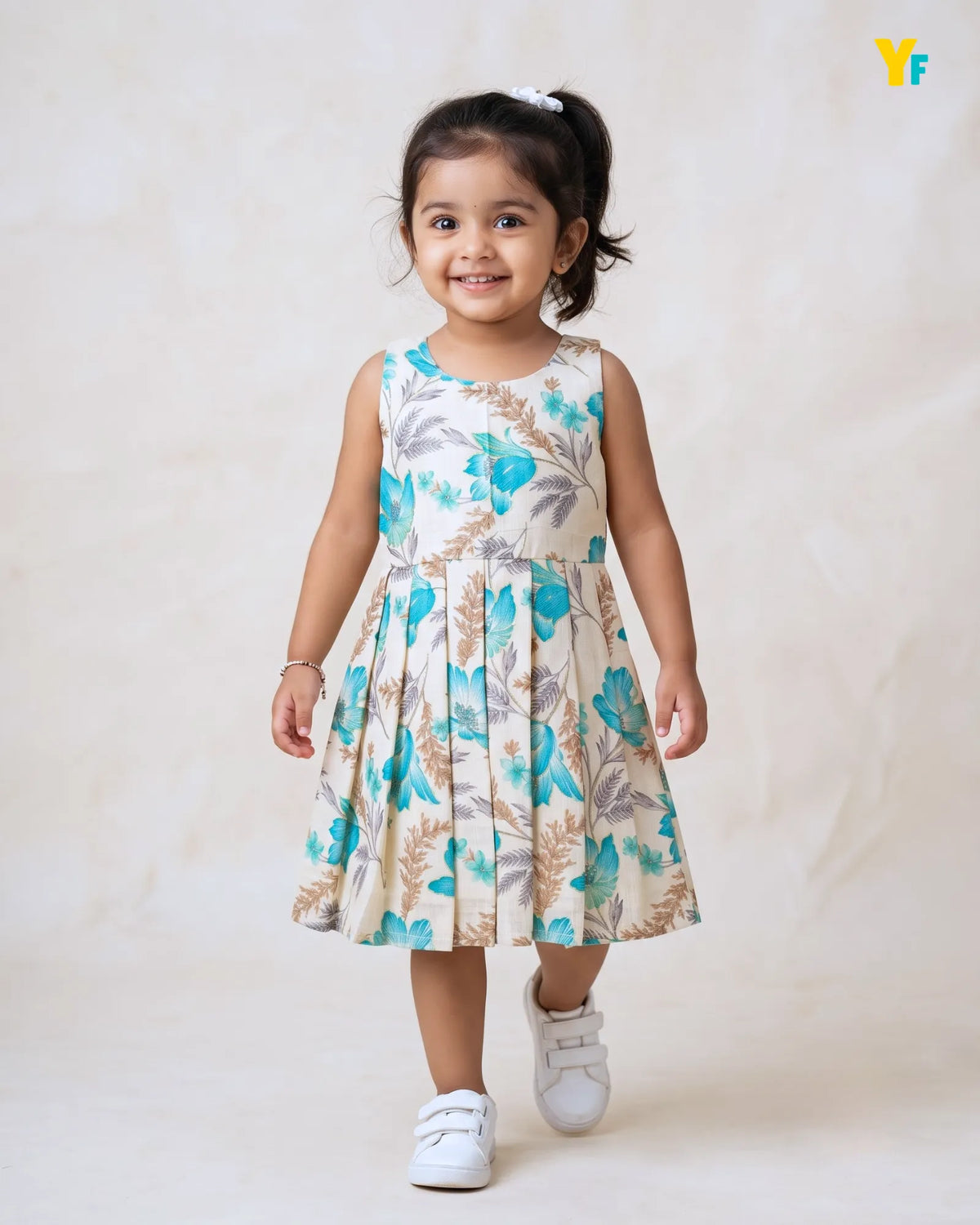 A young girl with dark hair in a ponytail is wearing a sleeveless, pleated A-line dress with a blue, green, and beige botanical pattern. She is walking forward on a light beige textured studio background, wearing white sneakers.