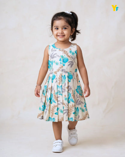 A young girl with dark hair in a ponytail is wearing a sleeveless, pleated A-line dress with a blue, green, and beige botanical pattern. She is walking forward on a light beige textured studio background, wearing white sneakers.