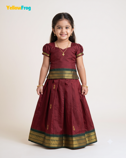A young girl modeling a traditional Indian maroon dress with gold and green borders, standing against a plain off-white background. The YellowFrog logo is visible in the top left corner.
