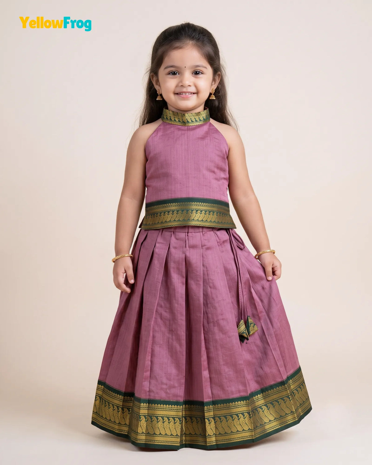 A young girl modeling a traditional Indian outfit consisting of a sleeveless dusty pink top and a matching pleated skirt with green and gold borders, set against a plain beige background.