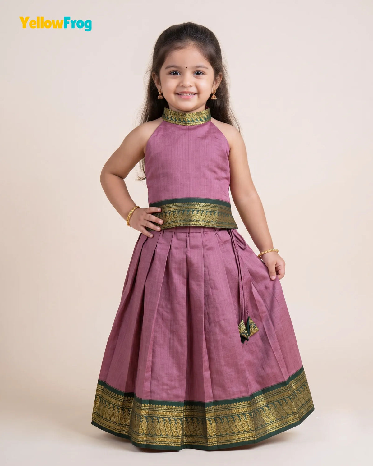 A young girl modeling a sleeveless dusty pink pleated traditional dress with green and gold borders, standing against a solid beige background. The YellowFrog logo is visible in the top left corner.
