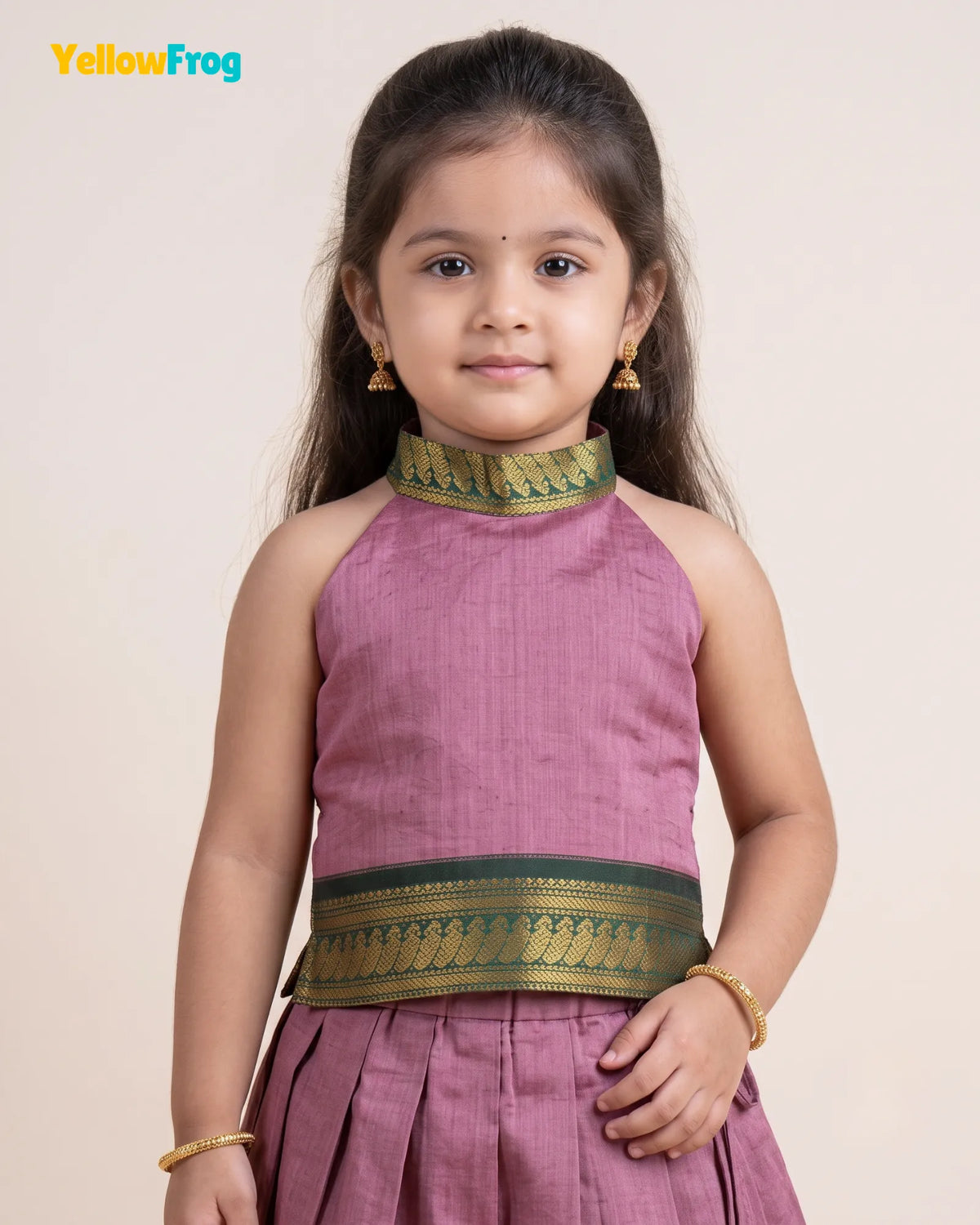 A young girl modeling a sleeveless dusty pink traditional Indian outfit with green and gold patterned borders at the neckline and waist, set against a plain beige background.