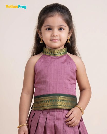 A young girl modeling a sleeveless dusty pink traditional Indian outfit with green and gold patterned borders at the neckline and waist, set against a plain beige background.