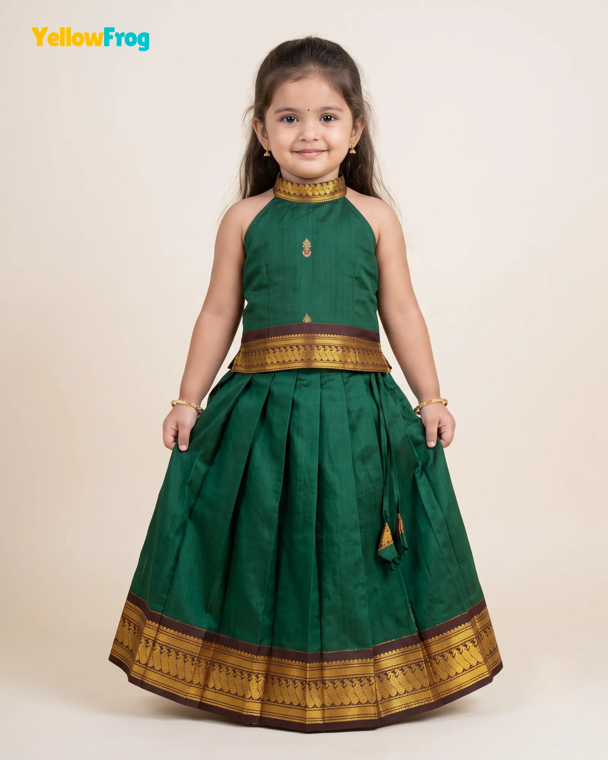 A young girl modeling a dark green traditional Indian dress with gold and brown borders, standing against a plain beige background. The YellowFrog logo is visible in the top left corner.