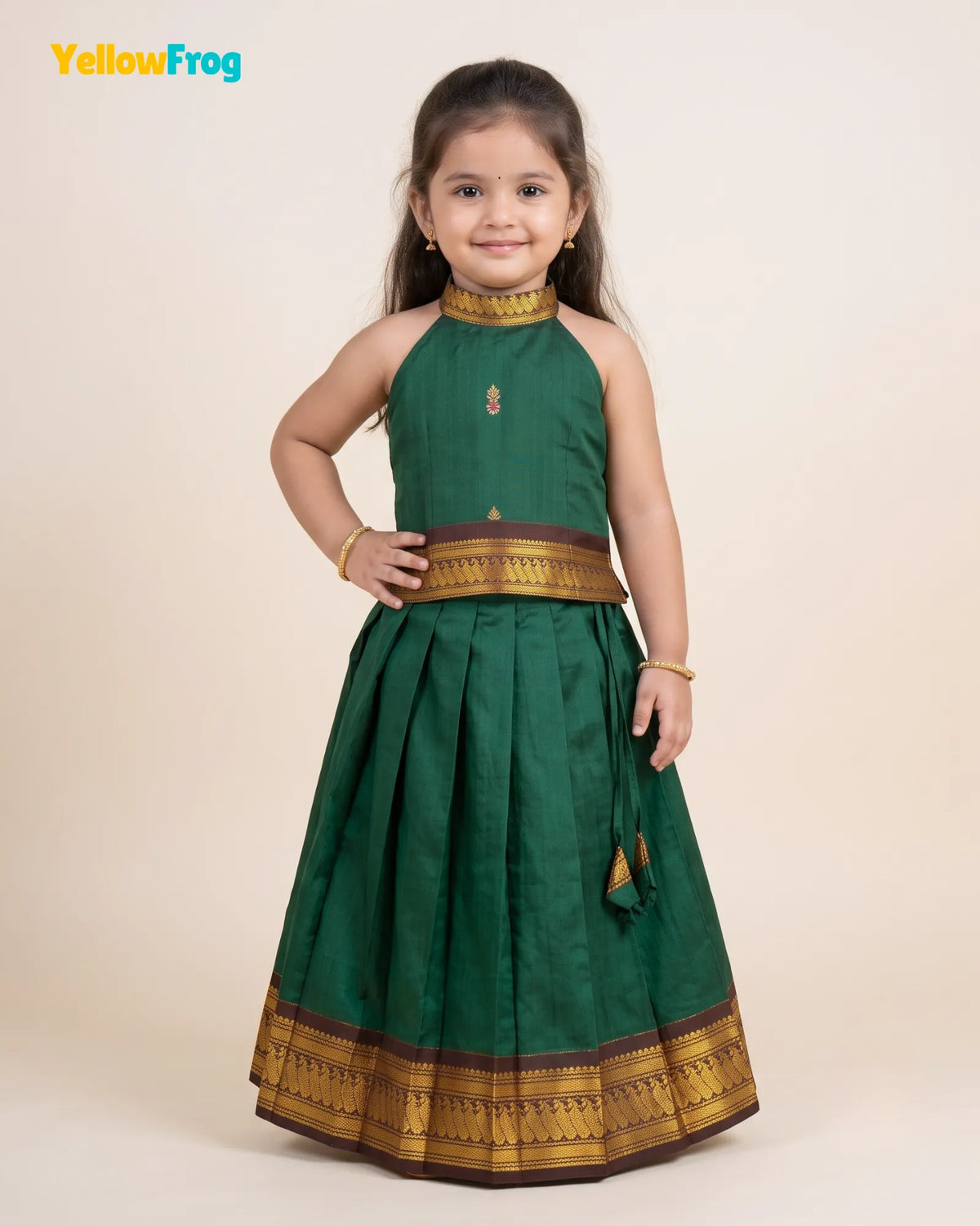 A young girl modeling a dark green sleeveless traditional dress with gold and maroon zari borders, set against a plain beige studio background. The YellowFrog brand logo is visible in the top left corner.