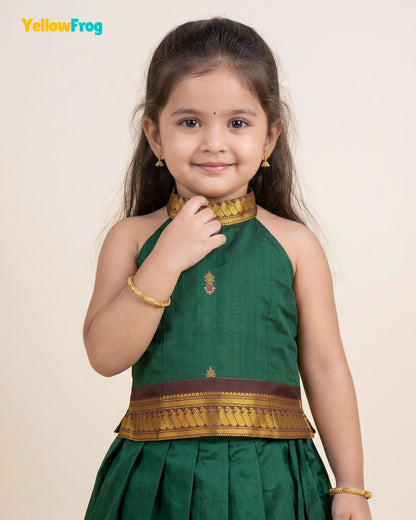 A young girl modeling a dark green traditional Indian dress with gold and brown zari embroidery, featuring a high neckline, halter design, and pleated skirt, set against a plain beige background.