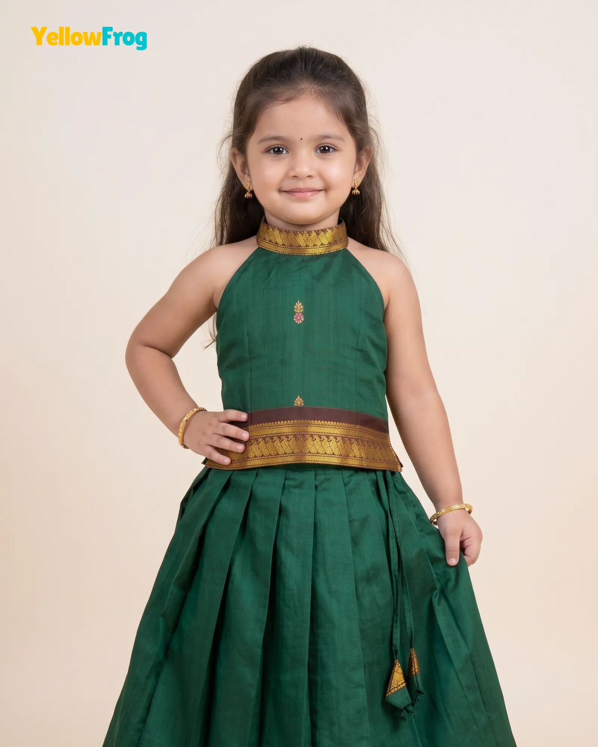 A young girl modeling a dark green traditional Indian dress with gold and brown decorative borders, set against a plain beige background. The YellowFrog logo is visible in the top left corner.