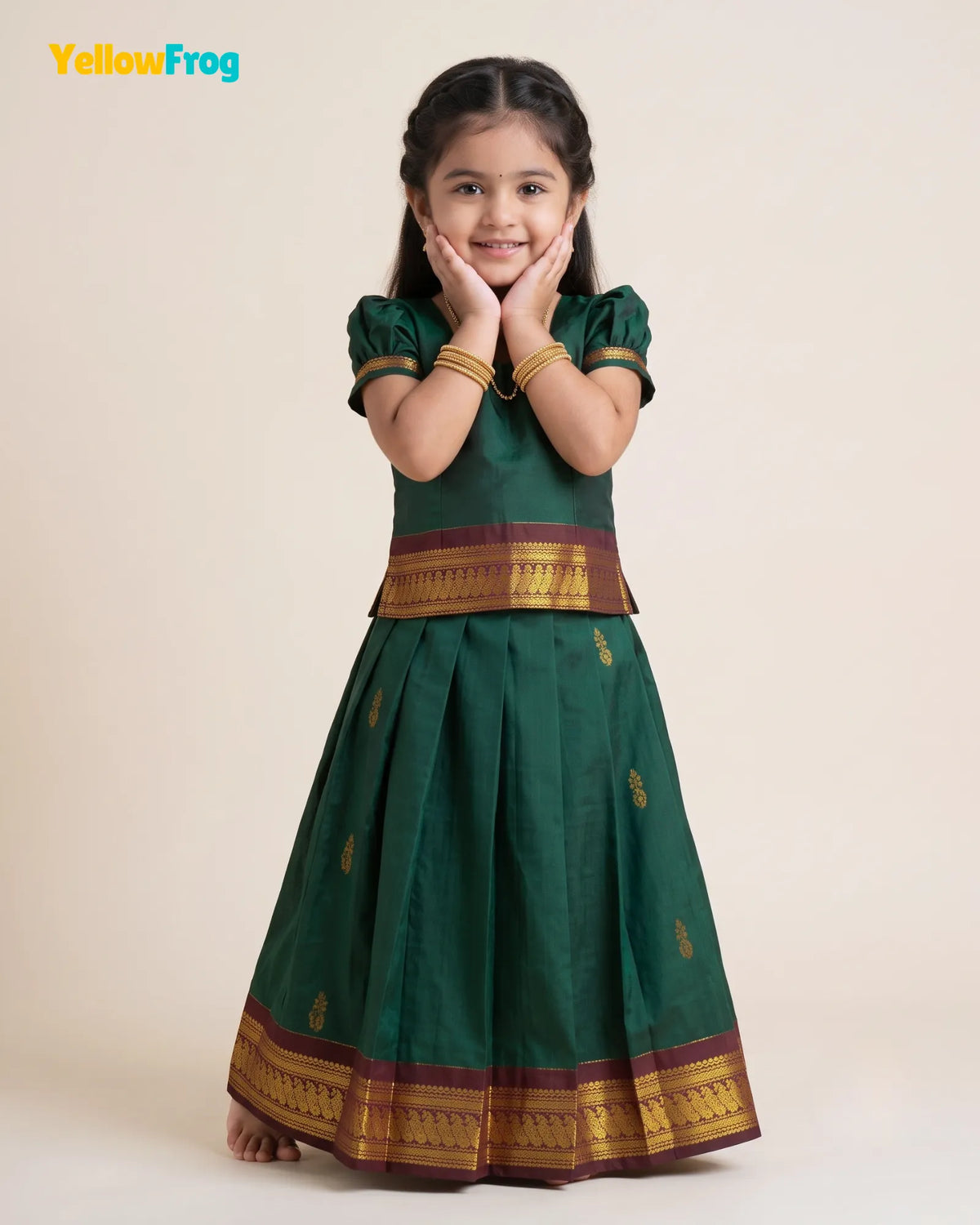 A young girl modeling a dark green traditional Indian outfit with gold and maroon borders, standing against a plain beige background. The YellowFrog logo is visible in the top left corner.