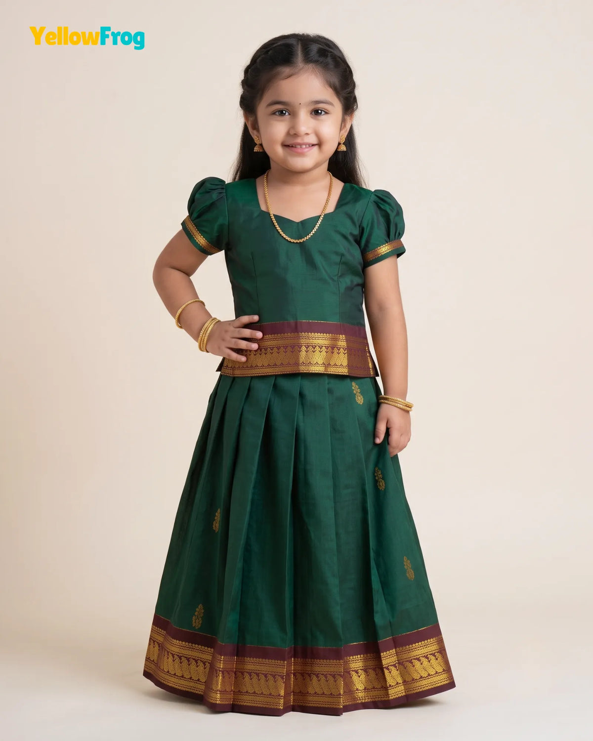 A young girl modeling a dark green traditional Indian dress with gold and maroon borders, standing against a plain beige studio background. The YellowFrog logo is visible in the top left corner.
