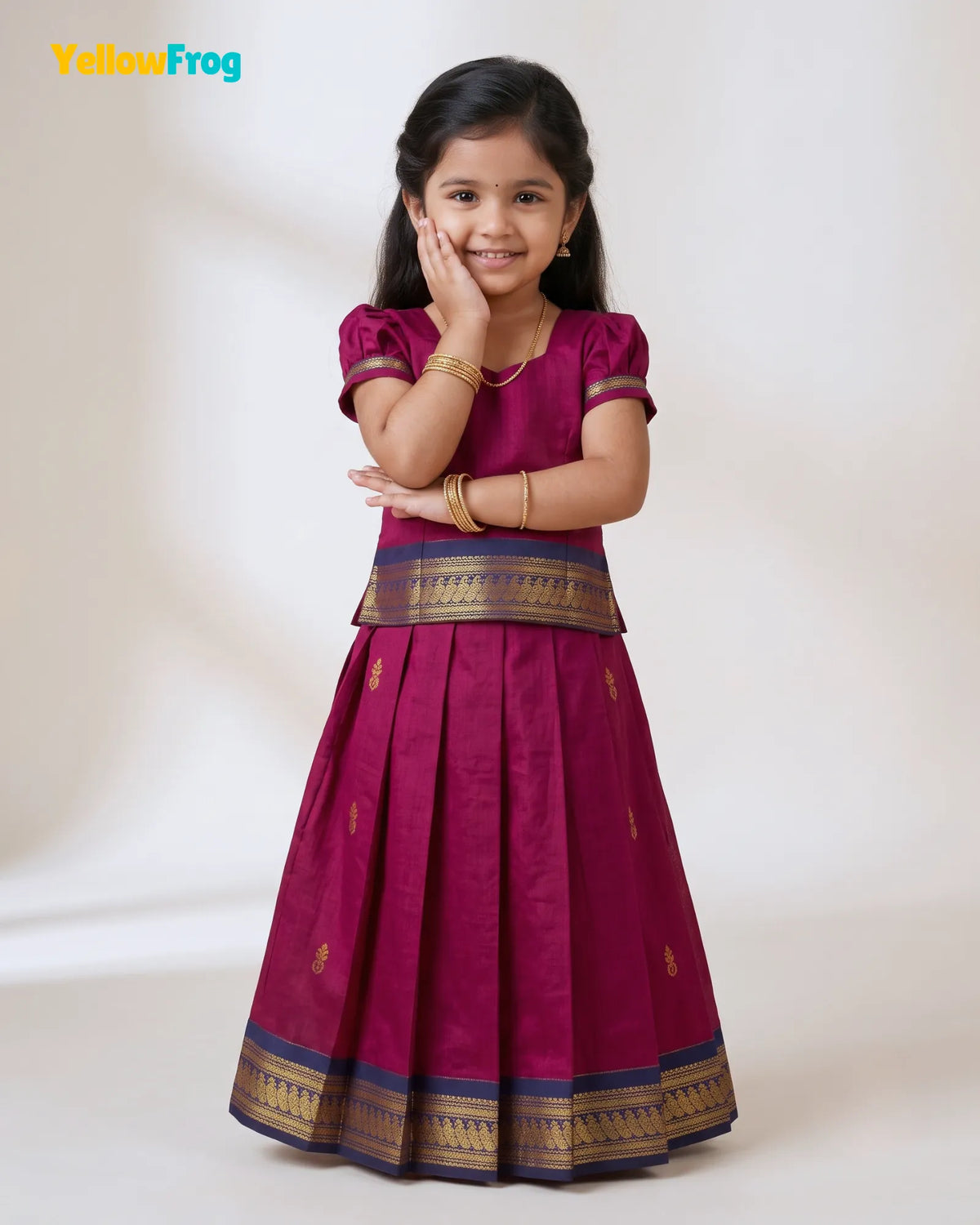A young girl modeling a traditional Indian outfit consisting of a magenta skirt with gold and navy borders, a matching magenta top, and gold jewelry, set against a plain cream background with a YellowFrog logo in the top left corner.