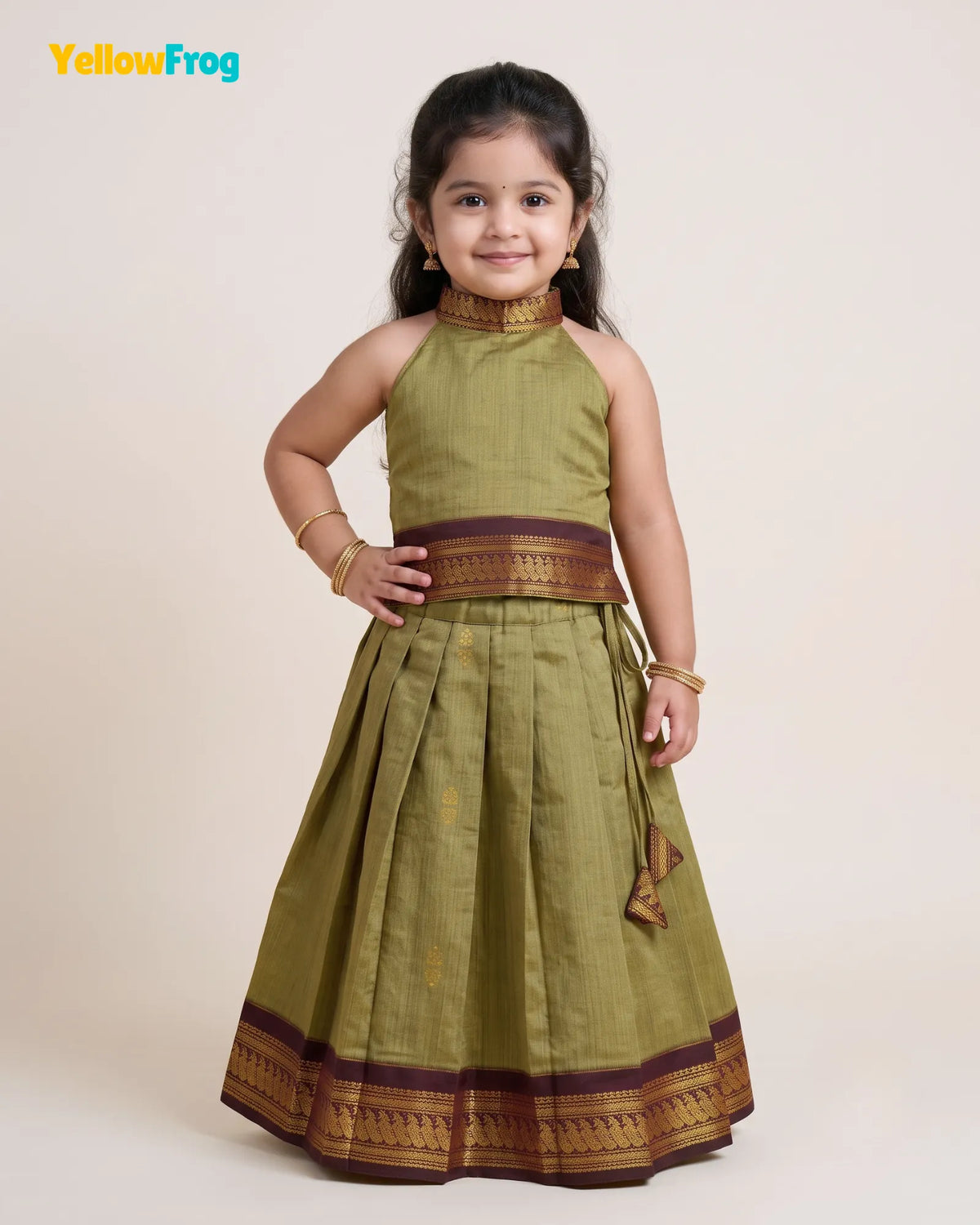 A young girl modeling a sleeveless olive green traditional dress with gold and brown borders, standing against a plain beige background. The YellowFrog logo is visible in the top left corner.