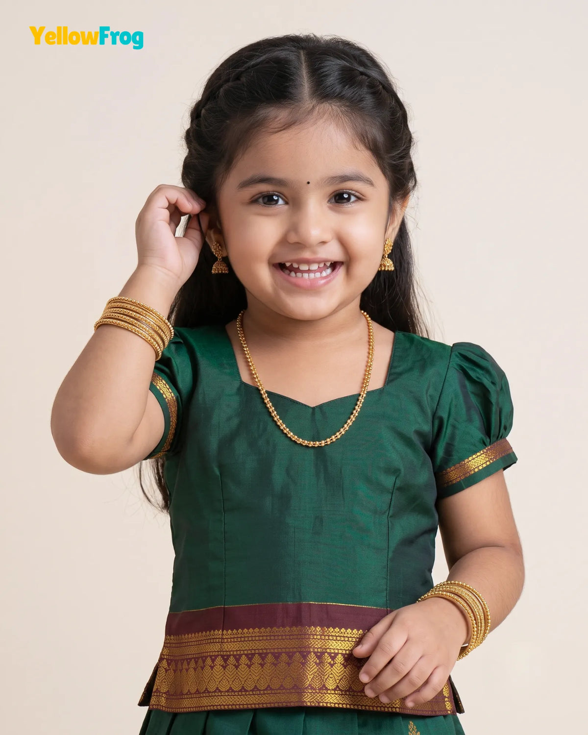 A young girl smiling and wearing a dark green traditional outfit with gold and maroon zari borders, along with gold jewelry, against a plain beige background. The YellowFrog logo is visible in the top left corner.