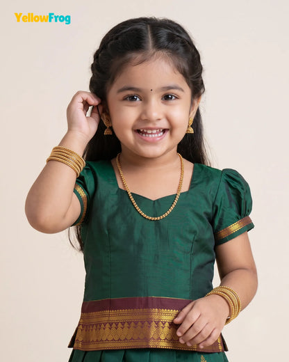 A young girl smiling and wearing a dark green traditional outfit with gold and maroon zari borders, along with gold jewelry, against a plain beige background. The YellowFrog logo is visible in the top left corner.