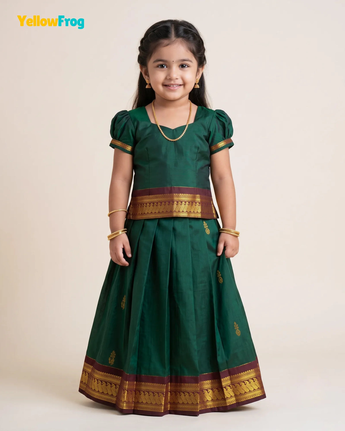 A young girl modeling a dark green traditional Indian dress with gold and maroon borders, standing against a plain beige background. The YellowFrog logo is visible in the top left corner.