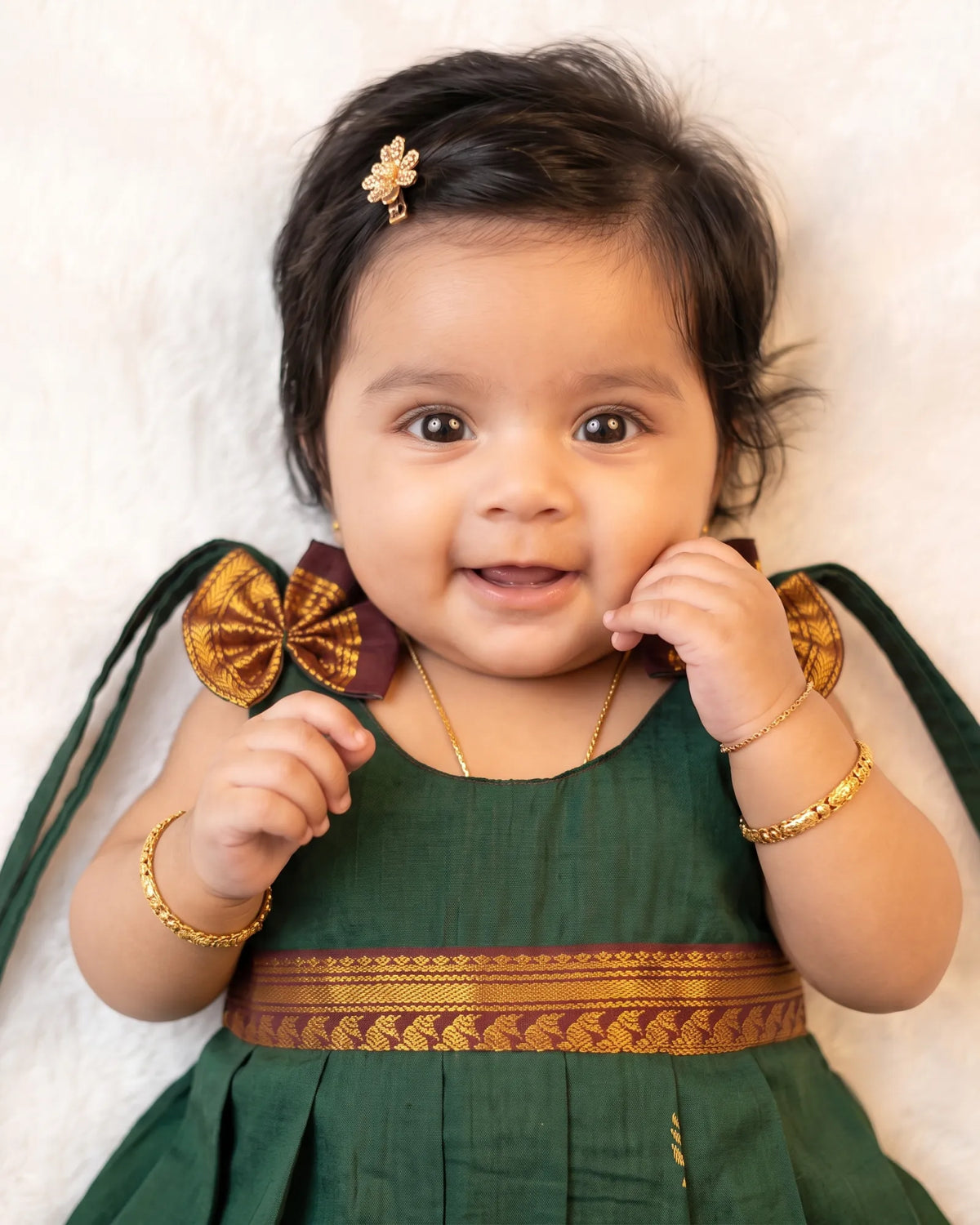 A smiling baby girl wearing a dark green traditional dress with gold and maroon borders, gold jewelry, and a small gold hair accessory, lying on a white textured background.