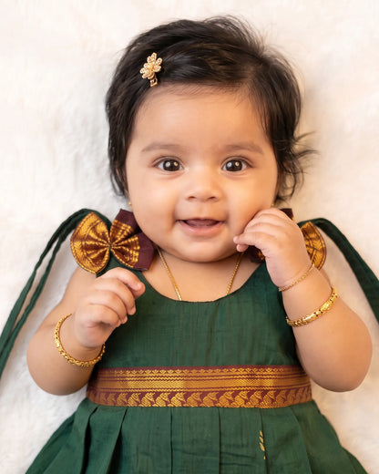 A smiling baby girl wearing a dark green traditional dress with gold and maroon borders, gold jewelry, and a small gold hair accessory, lying on a white textured background.