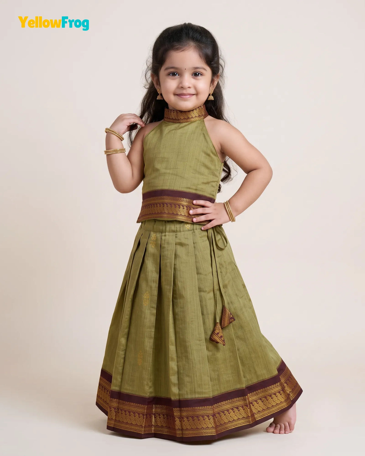 A young girl modeling a sleeveless olive green traditional dress with brown and gold zari borders, set against a plain beige studio background. The YellowFrog logo is visible in the top left corner.
