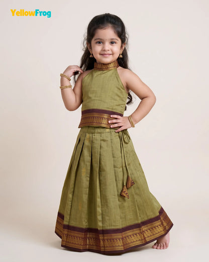 A young girl modeling a sleeveless olive green traditional dress with brown and gold zari borders, set against a plain beige studio background. The YellowFrog logo is visible in the top left corner.