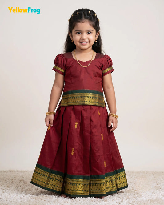 A young girl modeling a traditional Indian maroon salwar kameez outfit with gold and green borders, standing against a plain beige background.