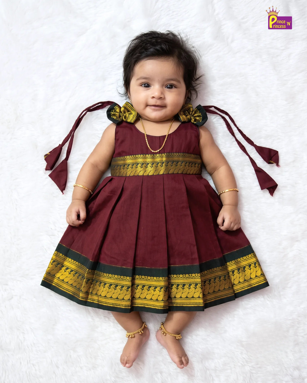 A baby girl wearing a maroon and gold traditional Indian dress, lying on a white textured blanket. The dress features a pleated skirt, a gold patterned waistband, and matching gold patterned shoulder bows with long ties.