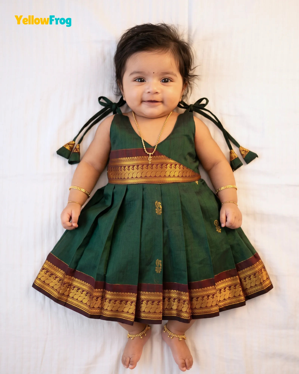 A baby girl wearing a dark green traditional Indian dress with gold and maroon borders, lying on a white sheet. The brand name 'YellowFrog' is visible in the top left corner.