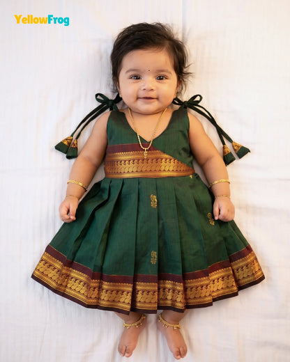A baby girl wearing a dark green traditional Indian dress with gold and maroon borders, lying on a white sheet. The brand name 'YellowFrog' is visible in the top left corner.
