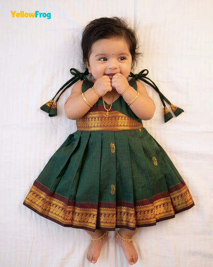 A baby girl wearing a dark green traditional Indian dress with gold and maroon borders, lying on a white sheet. The brand name 'YellowFrog' is visible in the top left corner.