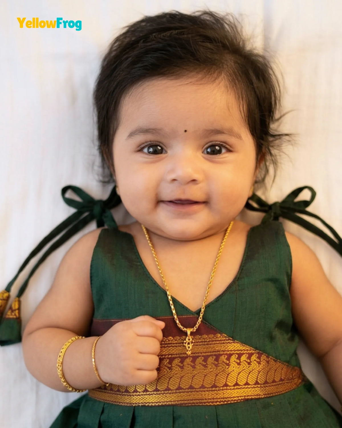 A smiling baby girl wearing a dark green traditional Indian dress with a gold embroidered waistband, a gold necklace, and a gold bracelet, lying on a white textured background. The 'YellowFrog' brand logo is visible in the top left corner.