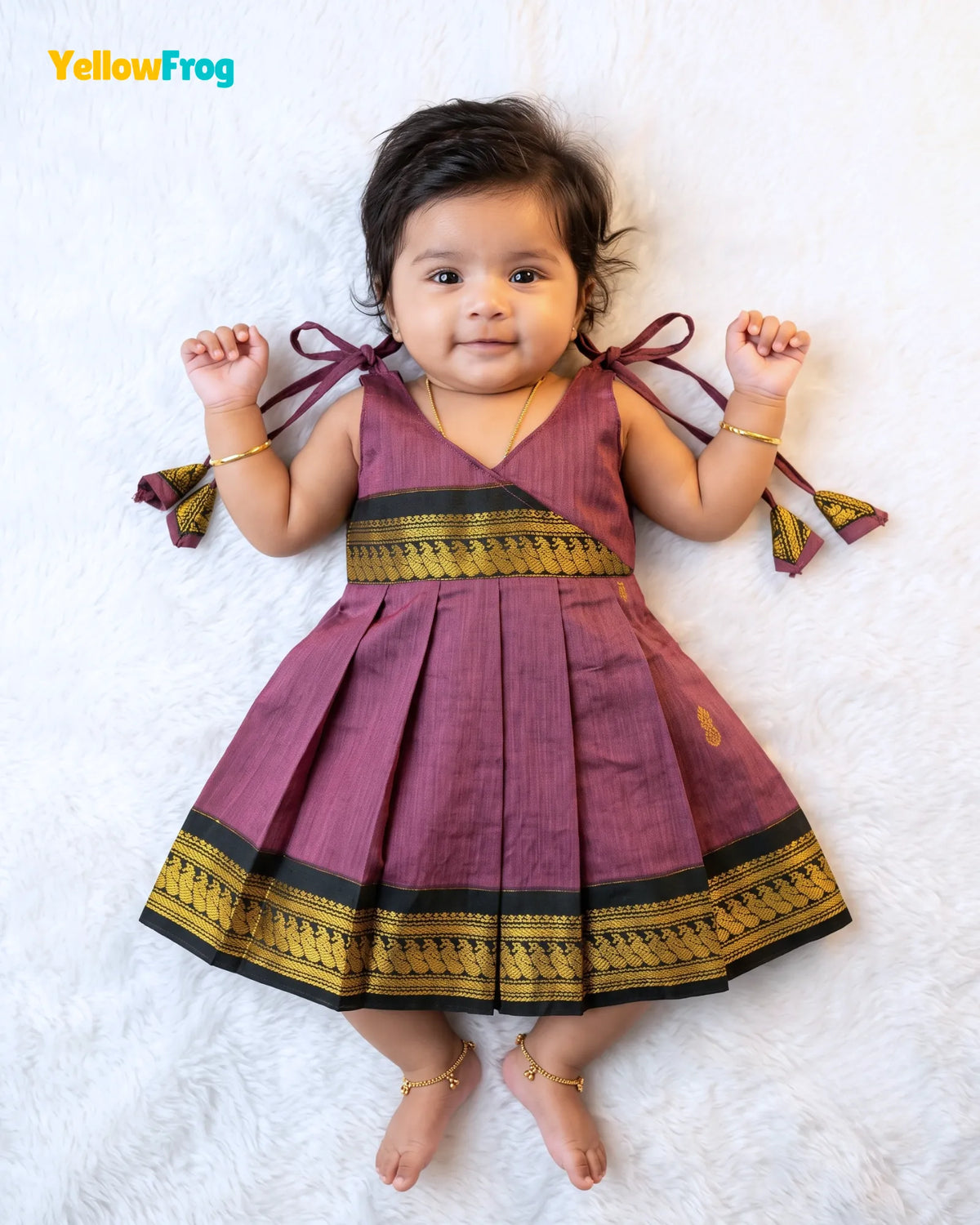 A baby girl lying on a white textured blanket wearing a sleeveless pleated dress in a dusty purple/magenta silk-like fabric with black and gold borders. The dress has tie-shoulder straps and a V-neckline.