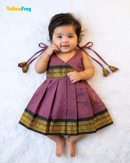 A toddler girl lying on a white faux fur rug wearing a pleated maroon dress with gold and black borders and tie details on the shoulders. The YellowFrog logo is visible in the top left corner.