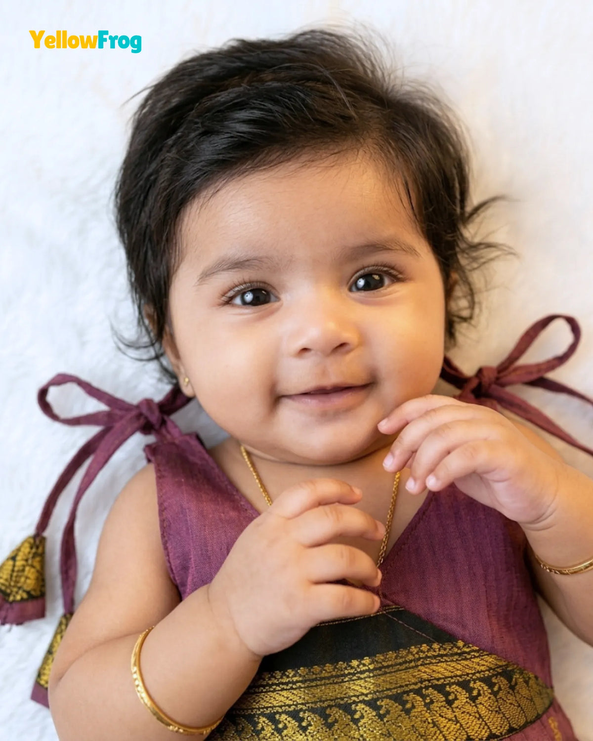 A close-up of a baby wearing a traditional Indian outfit in a deep purple and gold silk saree, lying on a white textured blanket. The brand name 'YellowFrog' is visible in the top left corner.