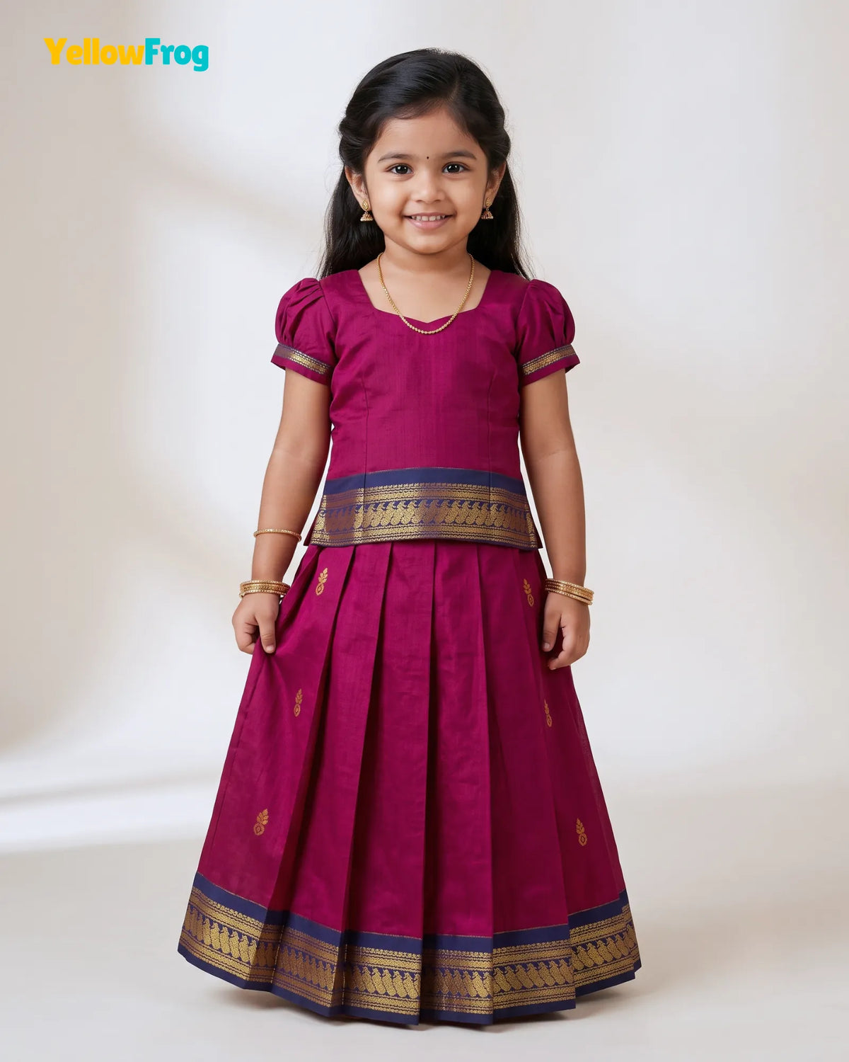 A young girl modeling a magenta pink traditional Indian dress with gold and navy borders, standing against a plain off-white background. The YellowFrog logo is visible in the top left corner.