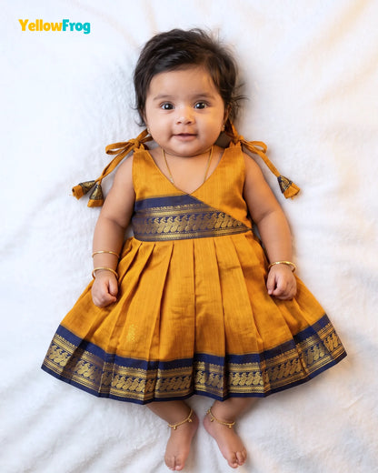 A baby girl wearing a mustard yellow and navy blue traditional dress with gold zari borders, lying on a white textured blanket. The YellowFrog brand logo is visible in the top left corner.
