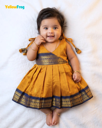 A smiling toddler girl lying on a white textured blanket, wearing a mustard yellow traditional dress with dark blue and gold borders and tie details on the shoulders.