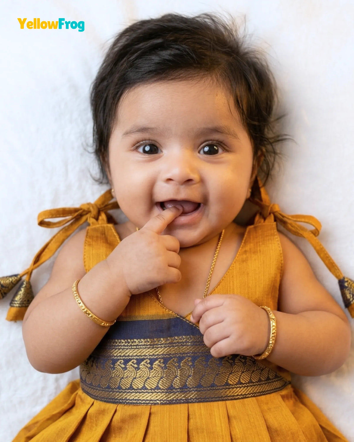 A baby wearing a mustard yellow traditional dress with a gold patterned waistband, lying on a white textured background. The brand name 'YellowFrog' is visible in the top left corner.