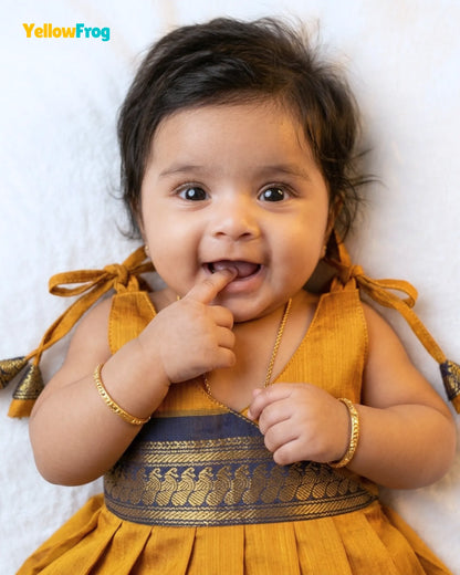 A baby wearing a mustard yellow traditional dress with a gold patterned waistband, lying on a white textured background. The brand name 'YellowFrog' is visible in the top left corner.