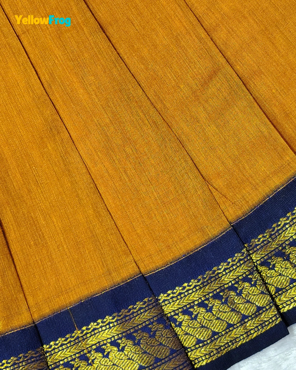 A close-up of a mustard yellow fabric, likely cotton or silk, showing its fine texture and a decorative border with a navy blue base and intricate gold woven pattern.